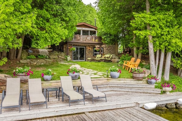 Waterfront deck with lounge chairs overlooking manicured lawn, stone cottage, and mature trees