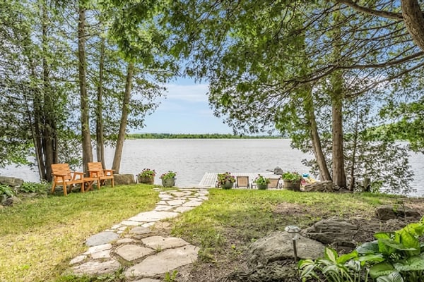 Lakeside yard with stone pathway, wooden Adirondack chairs, potted plants, and tall trees framing calm water view.