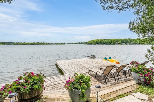Wooden dock on calm lake with lounge chairs, yellow kayak, pink flowering planters, and tree-lined shore in background